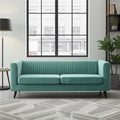 A mint green velvet loveseat sofa with vertical channel tufting on the backrest, shown in a modern living room setting with a gray patterned rug, a black floor lamp, and a window view.