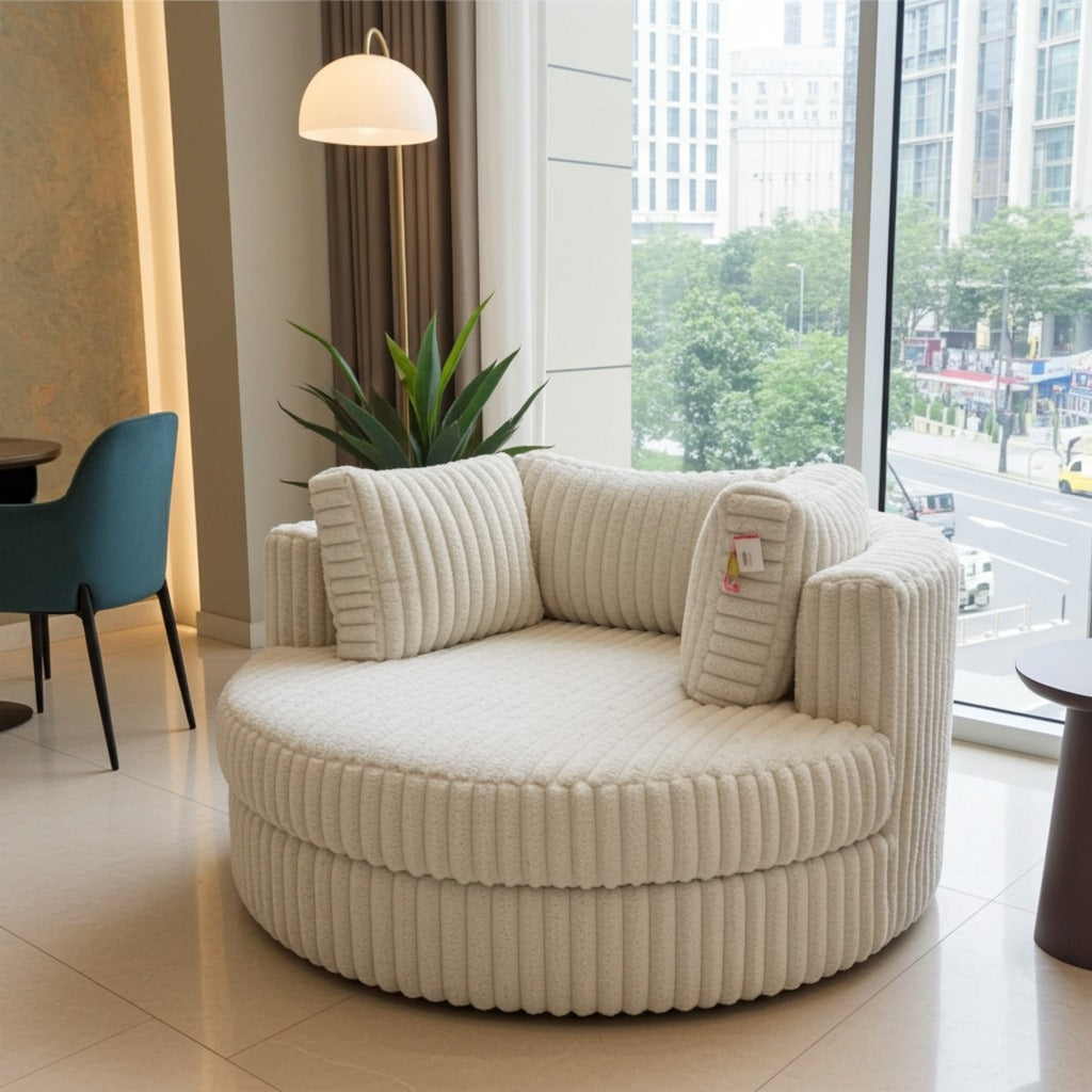 A round, cream-colored corduroy lounge chair with matching pillows, positioned in a modern living room setting with a large window showing a city view.