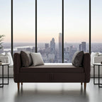 A modern brown upholstered bench with two throw pillows, positioned in front of large floor-to-ceiling windows overlooking a city skyline at sunset.
