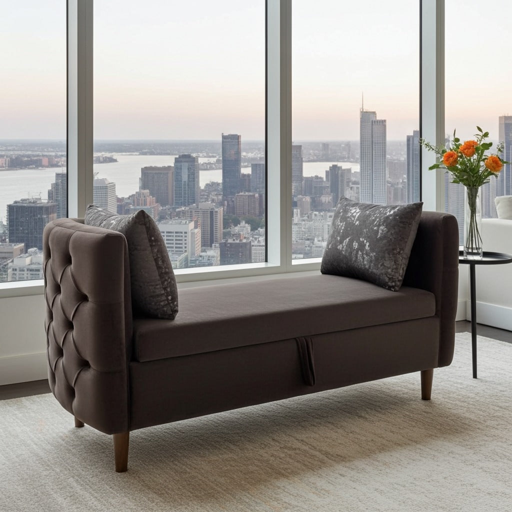 A dark brown upholstered bench with tufted arms and two dark throw pillows, positioned in front of large floor-to-ceiling windows overlooking a city skyline at sunset.