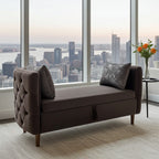 A dark brown upholstered bench with tufted arms and two dark throw pillows, positioned in front of large floor-to-ceiling windows overlooking a city skyline at sunset.