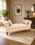 A cream-colored upholstered chaise lounge with rolled arms and wooden legs, styled in a warm, modern living room with a beige rug, potted plants, and wall art.