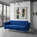 A modern blue upholstered loveseat with quilted chevron stitching and silver nailhead trim, positioned in a contemporary living room setting with a city view window, abstract wall art, and a floor lamp.