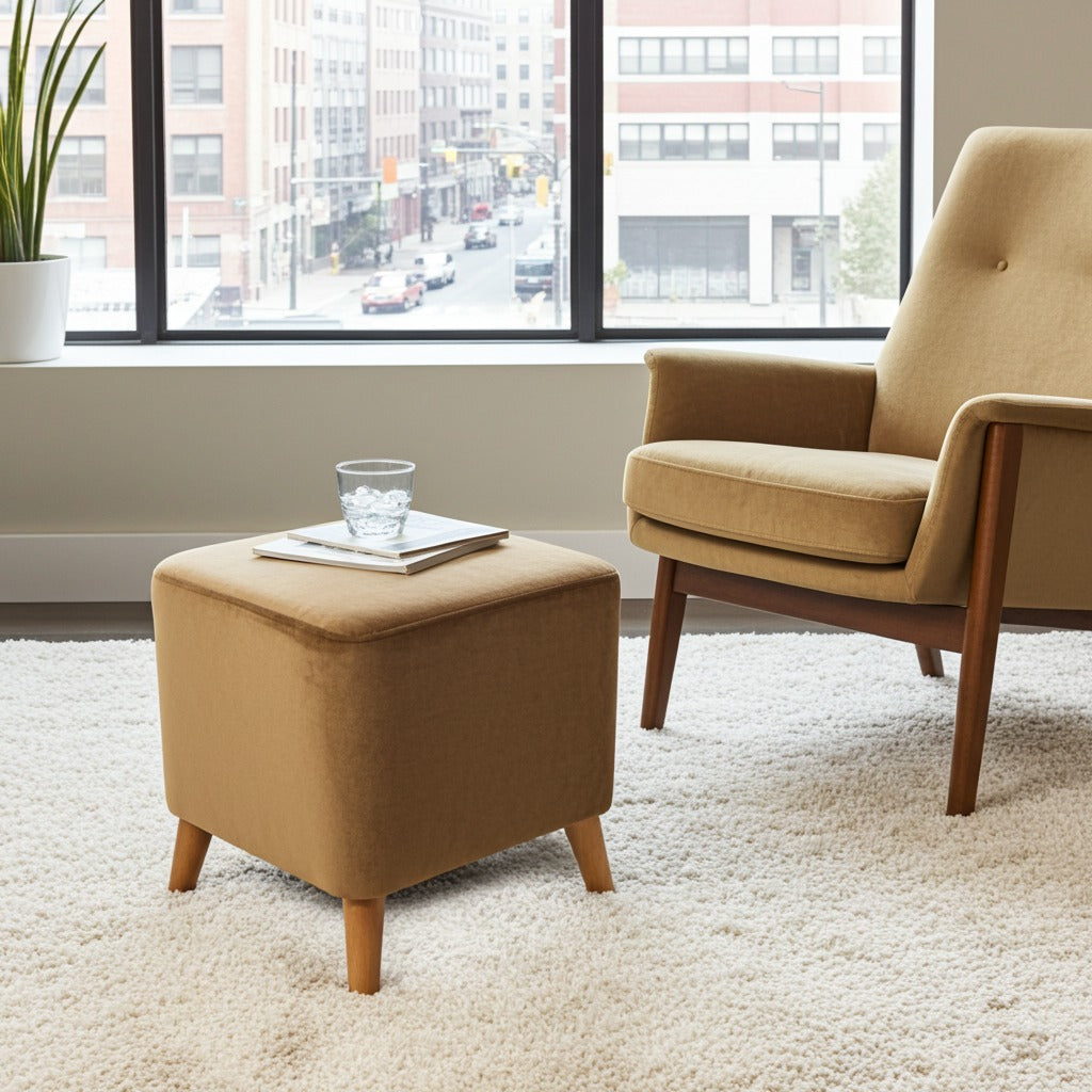 A tan upholstered ottoman with wooden legs sits on a white shag rug beside a matching armchair in a bright room with large windows showing a city street outside.