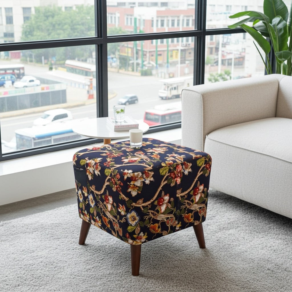 A square upholstered ottoman with a dark navy floral pattern and wooden legs, styled in a modern living room next to a white armchair and a small round table.