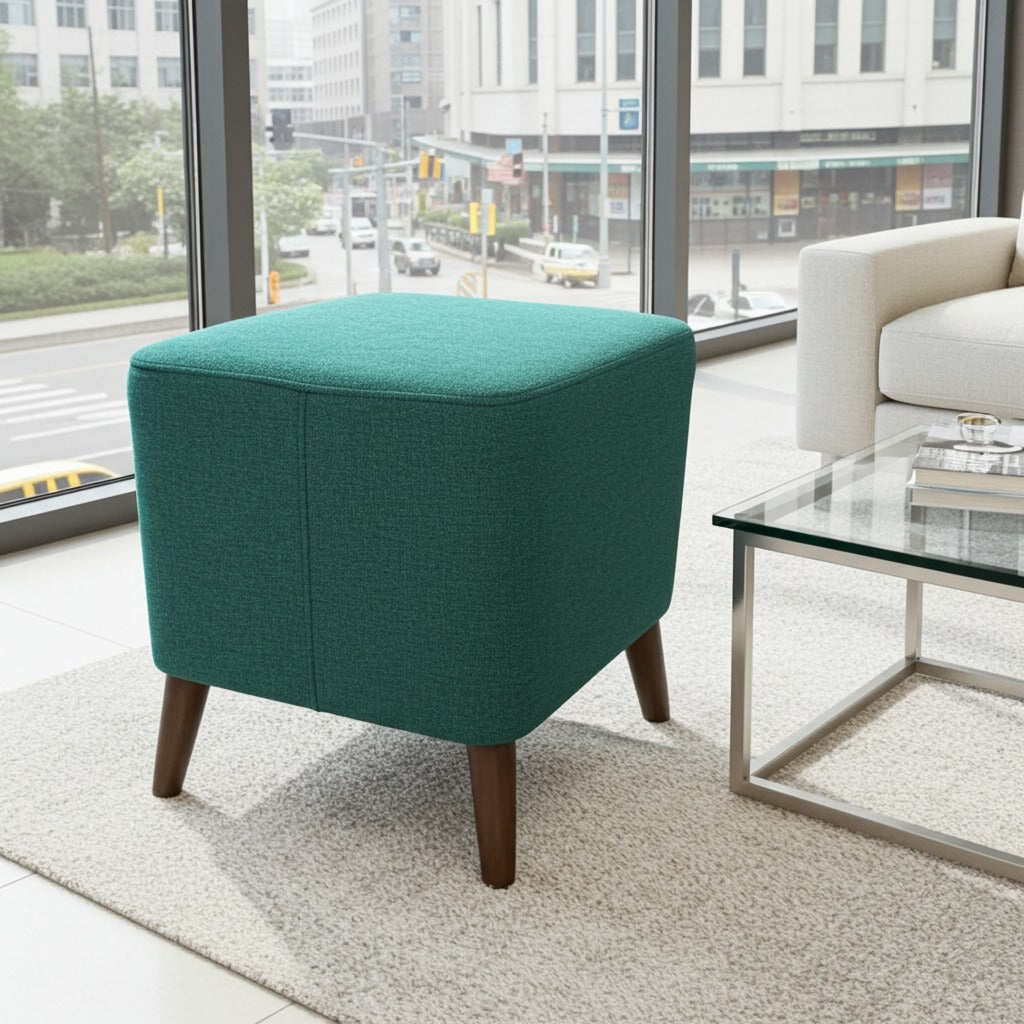 A square ottoman stool upholstered in textured teal-green fabric with tapered dark wood legs, positioned in a modern living room setting next to a glass coffee table and a white armchair.