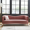 A curved upholstered sofa in dusty rose pink velvet with vertical channel tufting, gold metal legs, and a matching pink patterned accent pillow, set in a modern room with a large black-framed window, floor lamp, and patterned rug.
