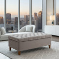 A tufted storage ottoman in a light taupe-beige fabric with wooden legs, positioned in a modern living room with a city skyline view through large floor-to-ceiling windows.