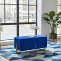 A bright blue upholstered rectangular ottoman with slim gold metal legs, placed on a blue and gray geometric area rug in a modern room with large windows, a potted plant, and a small side table.