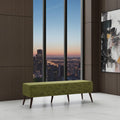An olive green upholstered bench with tapered dark wood legs, positioned in a modern interior with a city skyline view through large floor-to-ceiling windows.