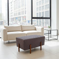 A modern living room setup featuring a cream-colored two-seater sofa and a dark gray quilted rectangular ottoman with wooden legs, set against a large window with a city view.