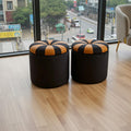 Two round black ottoman stools with striped tan and black cushioned lids, placed on a light wood floor in front of a large window overlooking a city street.