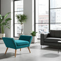 A modern teal upholstered accent chair with wooden legs and silver nailhead trim, shown in a bright contemporary living room next to a black sofa and large windows.