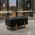 A modern black upholstered ottoman with four tapered wooden legs, positioned on a patterned rug in a contemporary office setting with a city skyline view.