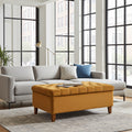 A modern living room scene featuring a light gray sectional sofa with accent pillows and a prominent mustard yellow tufted storage ottoman in the foreground, set on a patterned rug in front of large black-framed windows.