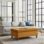 A modern living room scene featuring a light gray sectional sofa with accent pillows and a prominent mustard yellow tufted storage ottoman in the foreground, set on a patterned rug in front of large black-framed windows.
