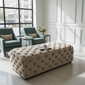 A large tufted ottoman bench in a warm beige fabric, styled in a bright, modern living room with two teal armchairs and a large window.