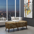 A leopard-print upholstered rectangular ottoman with dark wooden legs in a modern living room setting, positioned in front of a white sofa with a coffee table, with a city skyline visible through large windows and an abstract painting on the wall.