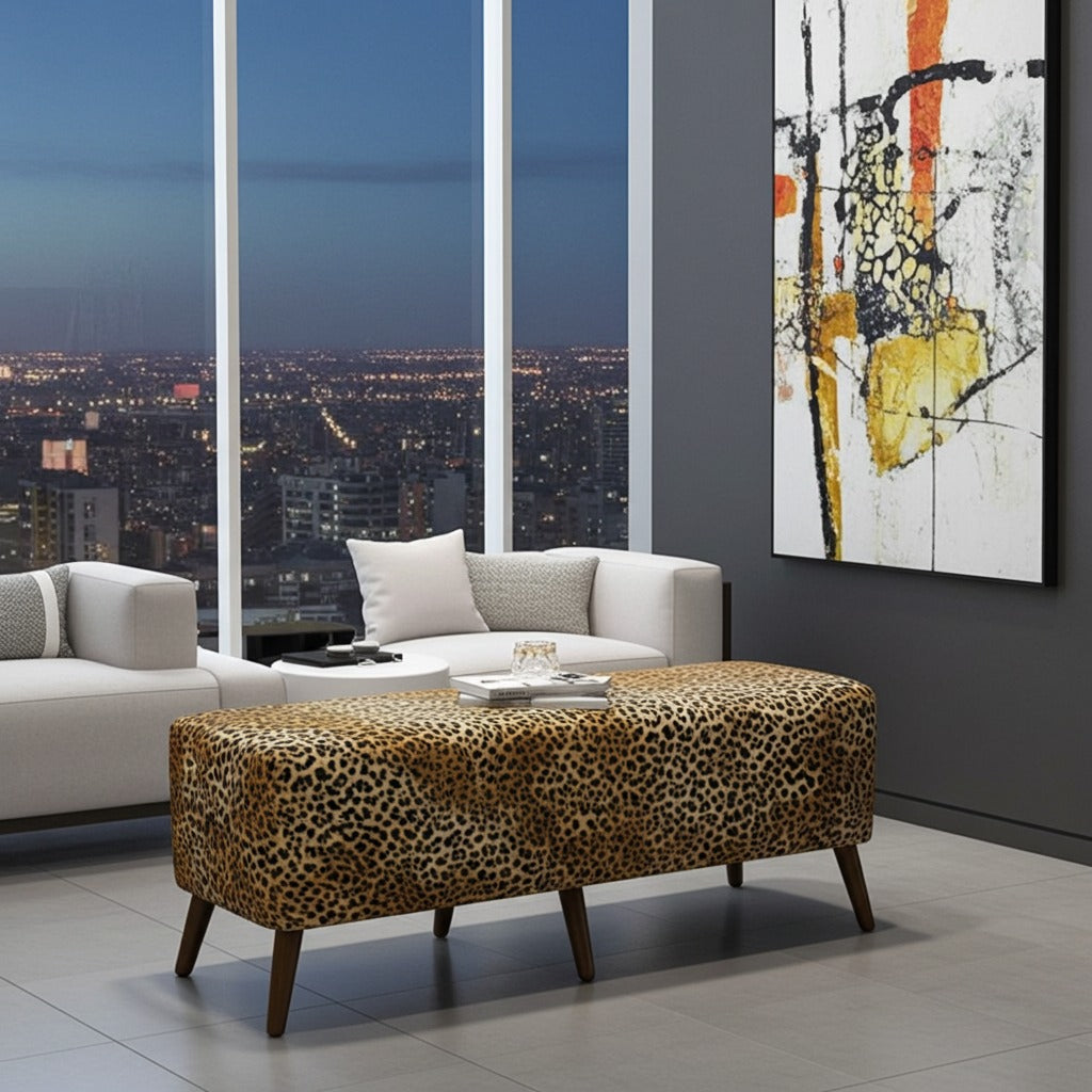 A leopard-print upholstered rectangular ottoman with dark wooden legs in a modern living room setting, positioned in front of a white sofa with a coffee table, with a city skyline visible through large windows and an abstract painting on the wall.
