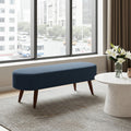 A modern navy blue upholstered bench with tapered wooden legs, positioned in a bright living room next to a marble-topped coffee table with white flowers.