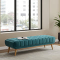 A teal tufted velvet bench with light wood legs, styled in a modern room with a large window showing an outdoor city view.