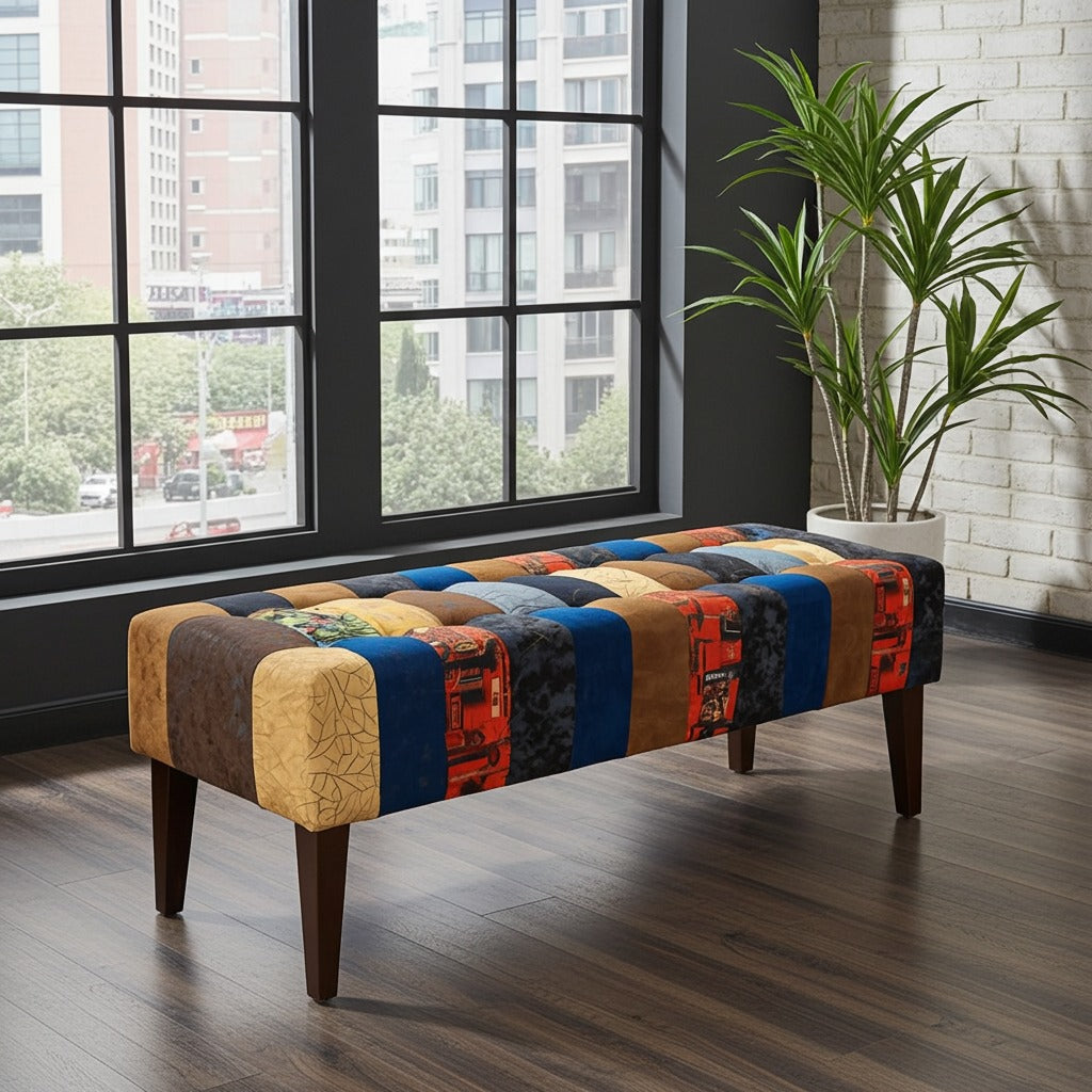 A rectangular tufted bench with a colorful patchwork fabric upholstery and dark wooden legs, positioned in a modern room with a large window, dark wood floor, and a potted plant.