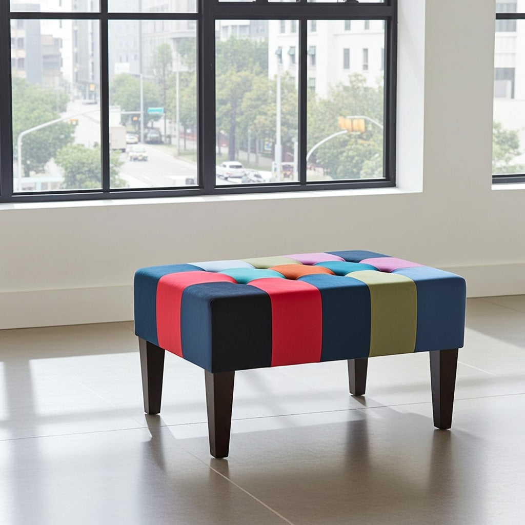 A square tufted ottoman with colorful patchwork upholstery and dark wooden legs, positioned in a bright room with large windows showing an outdoor view.