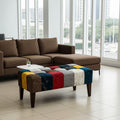 A modern living room setup featuring a brown sectional sofa and a low tufted ottoman upholstered in a colorful patchwork of wood-grain and fabric panels in blue, red, yellow, gray, and brown. The room has large floor-to-ceiling windows with a city view.