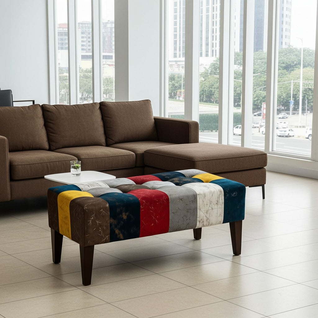 A modern living room setup featuring a brown sectional sofa and a low tufted ottoman upholstered in a colorful patchwork of wood-grain and fabric panels in blue, red, yellow, gray, and brown. The room has large floor-to-ceiling windows with a city view.