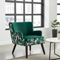 A modern accent chair with a deep green velvet back and seat, upholstered in a green and white tropical leaf pattern on the sides and backrest, with dark wooden legs. It is shown in a bright, contemporary room next to a white side table.