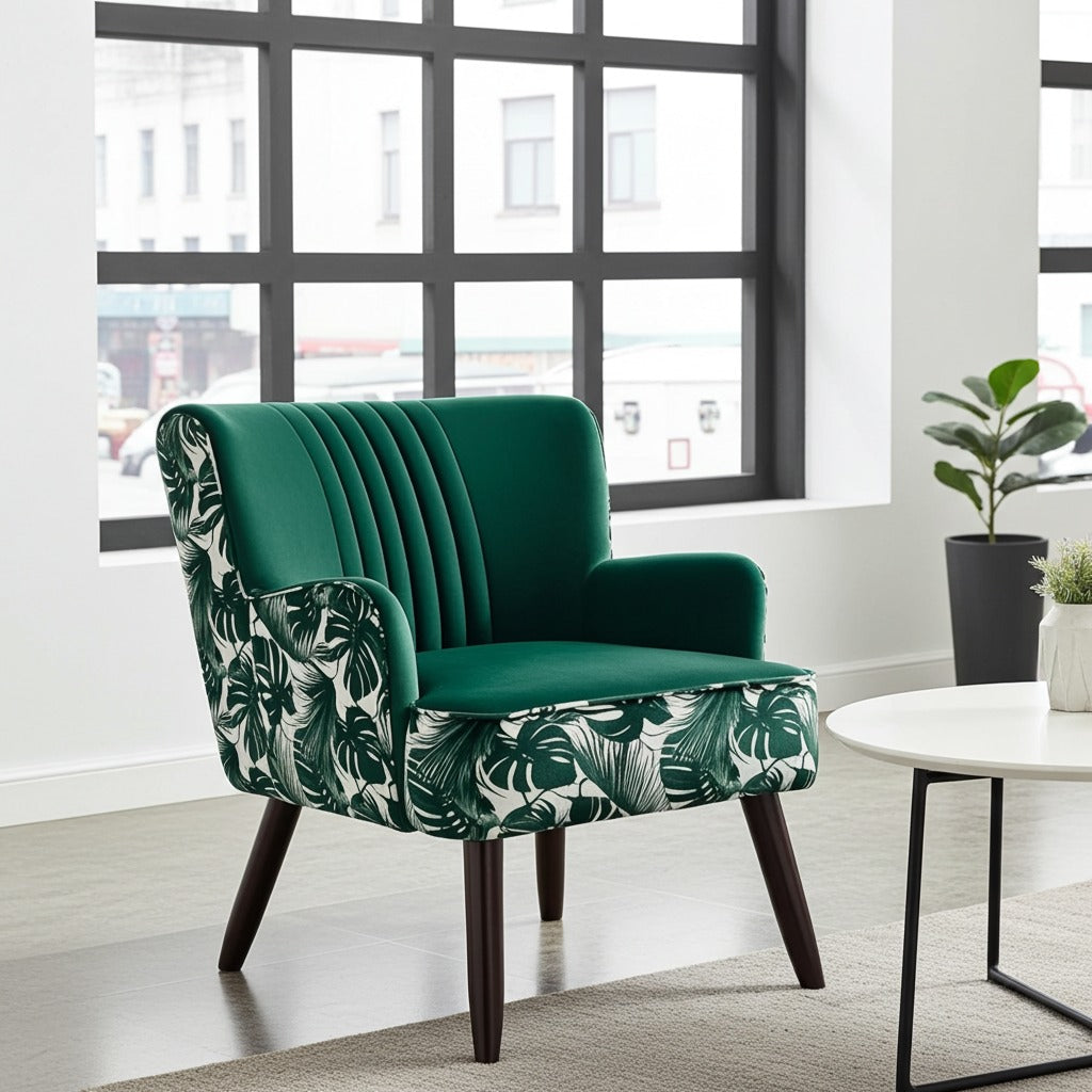 A modern accent chair with a deep green velvet back and seat, upholstered in a green and white tropical leaf pattern on the sides and backrest, with dark wooden legs. It is shown in a bright, contemporary room next to a white side table.