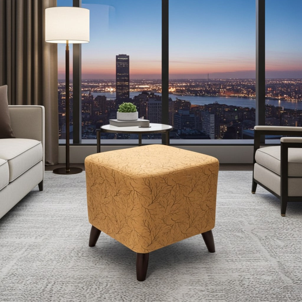 A mustard yellow upholstered ottoman with a subtle leaf pattern and dark wooden legs, styled in a modern living room with a city skyline view at dusk.