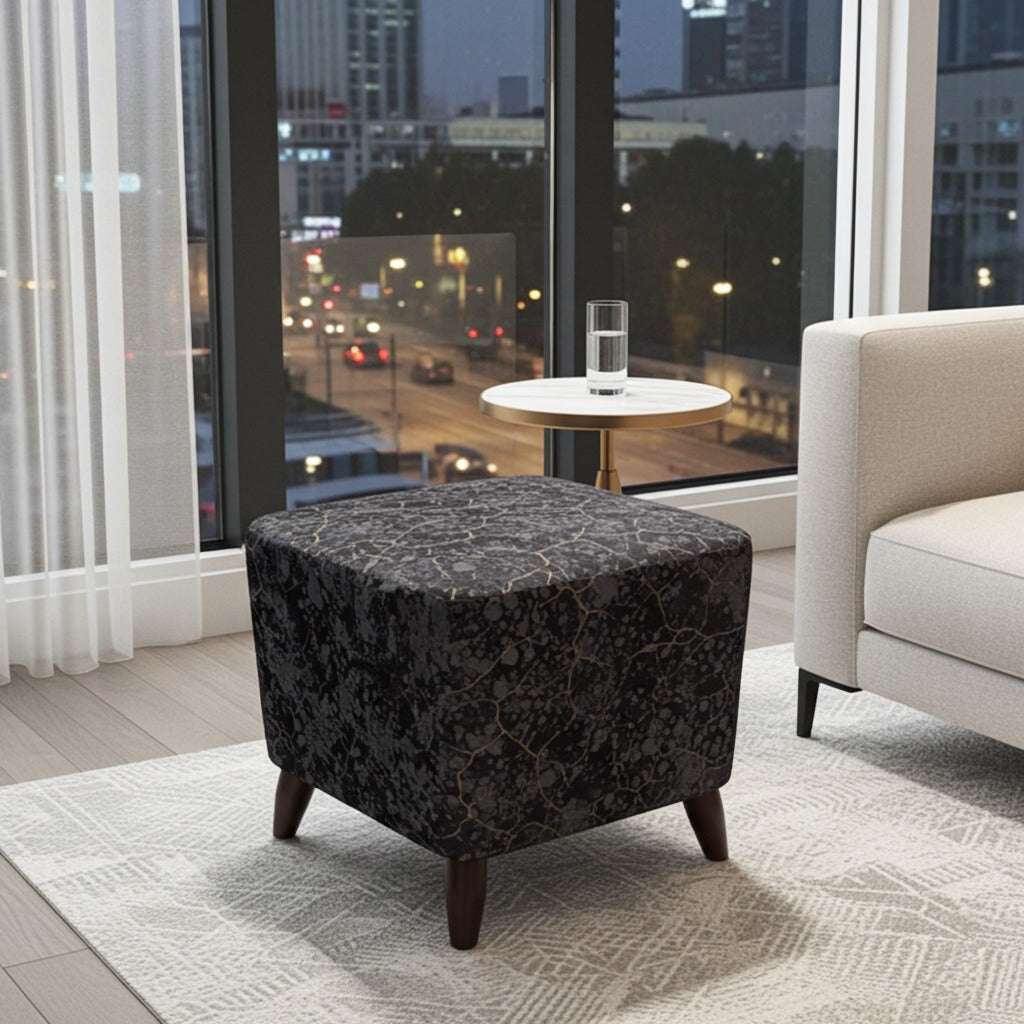 A square upholstered ottoman with a dark floral patterned fabric and tapered dark wooden legs, shown in a modern living room setting with a window view of a city street at dusk.