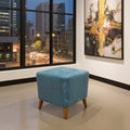A square ottoman stool with a distressed turquoise blue upholstery and light wood legs, positioned in a modern room with a large window showing a city street at dusk.