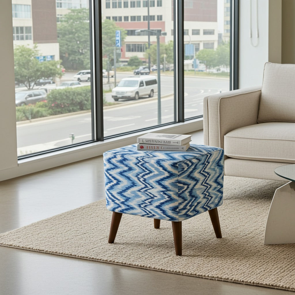 A blue and white patterned ottoman with dark wood legs sits on a beige rug in a modern living room setting with a large window view.