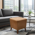 A square upholstered ottoman in a warm camel-brown fabric with tapered wooden legs, positioned in a modern living room next to a gray sofa and a small side table.