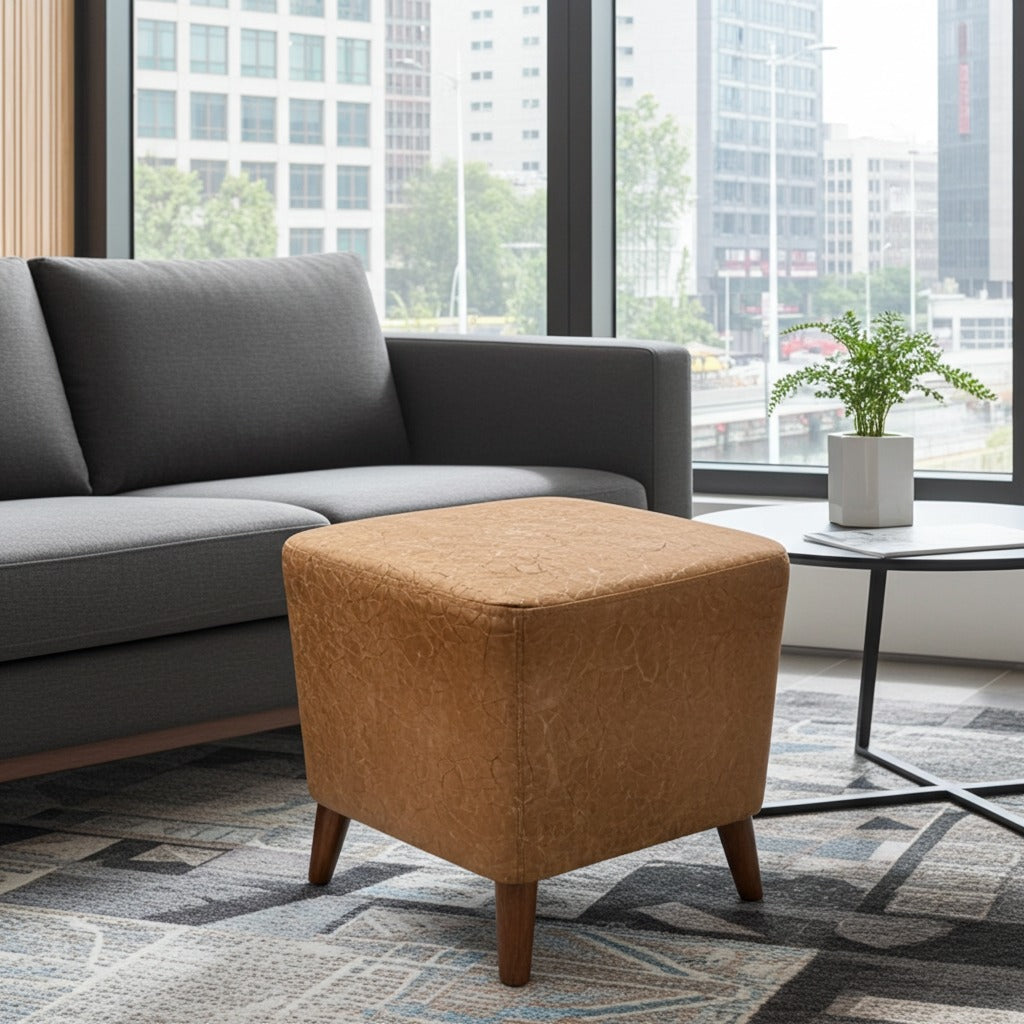 A square upholstered ottoman in a warm camel-brown fabric with tapered wooden legs, positioned in a modern living room next to a gray sofa and a small side table.