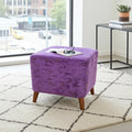 A square purple velvet ottoman with tapered wooden legs sits on a white shag rug in a bright room. An open book and glasses rest on top of the ottoman, with a black side table and window visible in the background.
