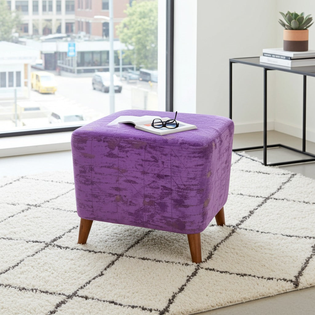 A square purple velvet ottoman with tapered wooden legs sits on a white shag rug in a bright room. An open book and glasses rest on top of the ottoman, with a black side table and window visible in the background.
