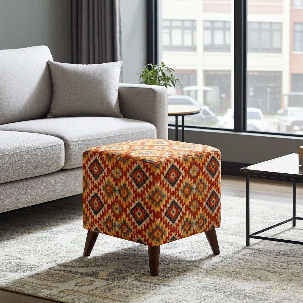 A square ottoman with a colorful geometric Southwestern-style pattern in orange, red, yellow, and blue, supported by four dark wood legs, shown in a modern living room setting next to a light gray sofa.