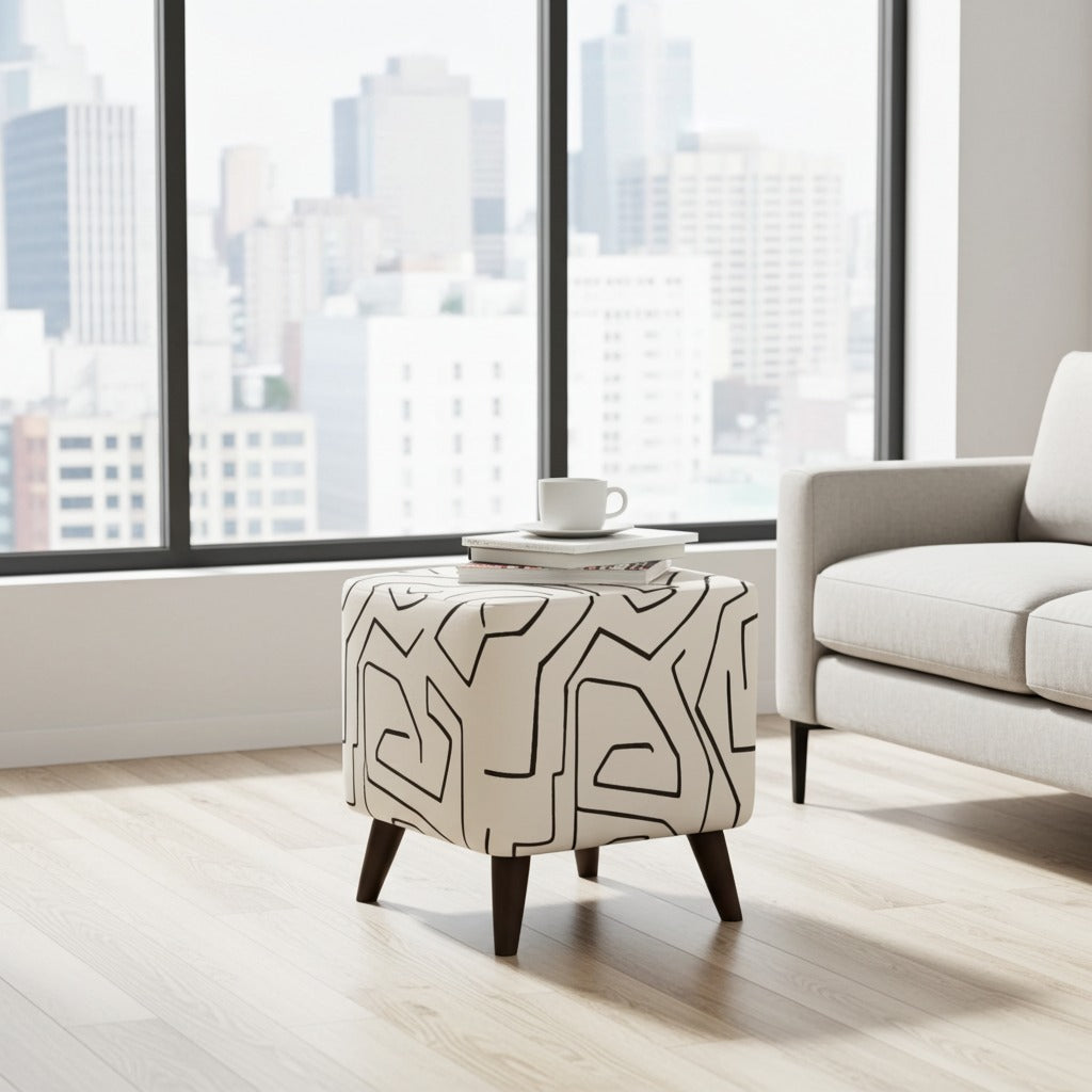 A square ottoman with dark legs and a cream upholstery covered in a black abstract line pattern, styled in a modern living room with a city view window and a light gray sofa.