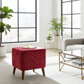 A square red upholstered ottoman with a subtle gold branch pattern and tapered wooden legs, styled in a bright, modern living room next to a glass coffee table and potted plants.