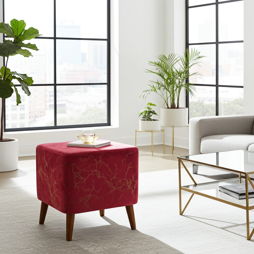 A square red upholstered ottoman with a subtle gold branch pattern and tapered wooden legs, styled in a bright, modern living room next to a glass coffee table and potted plants.