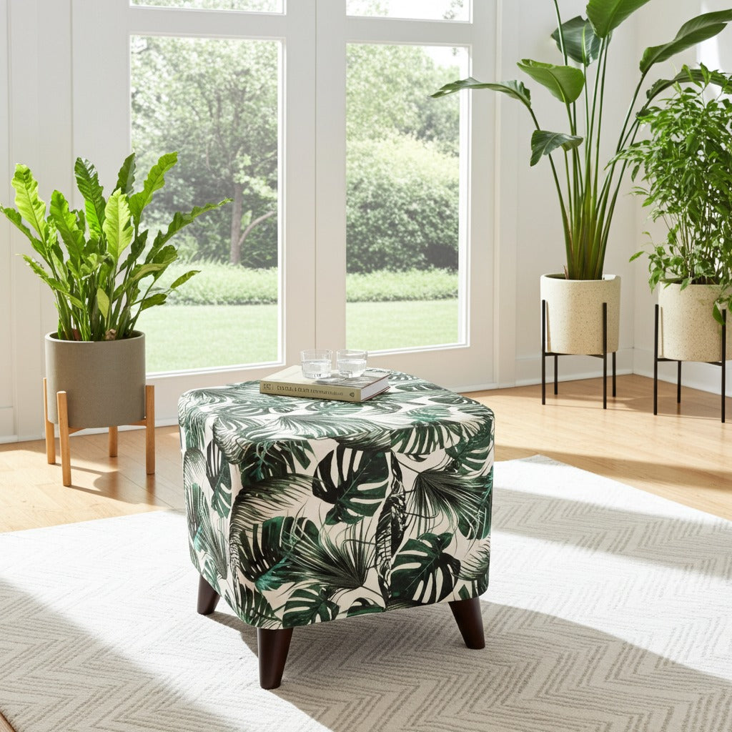 A square ottoman stool with a green and white tropical monstera leaf print, styled in a bright room with potted plants and large windows.