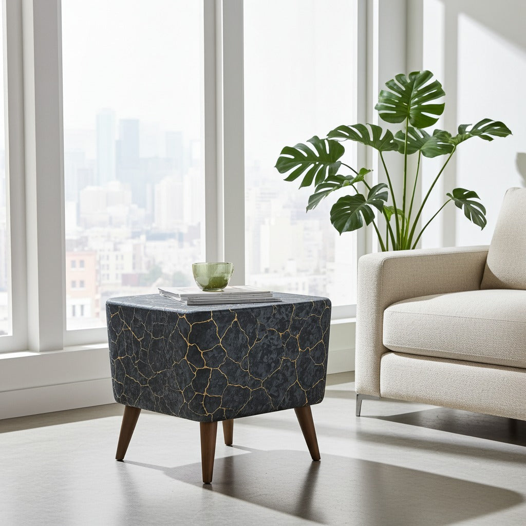A square ottoman with a dark gray and black marble-like surface featuring gold veins and four tapered wooden legs, styled in a bright modern room next to a beige armchair and a potted green plant.