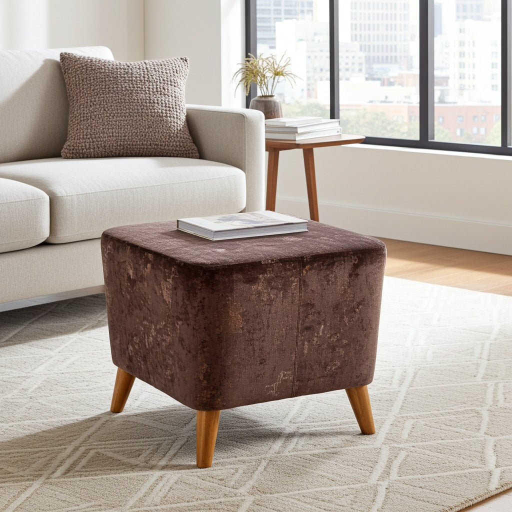 A square ottoman with a textured dark brown velvet-like fabric and tapered wooden legs, styled in a bright living room next to a white sofa.
