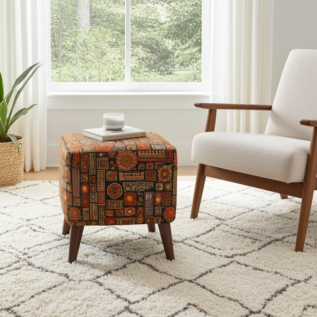 A square ottoman with a colorful geometric patchwork pattern in orange, brown, and red, styled in a bright living room next to a white armchair and a window.