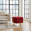 A square ottoman in deep red velvet with slim gold metal legs, styled in a modern living room next to a beige sofa and a glass coffee table.