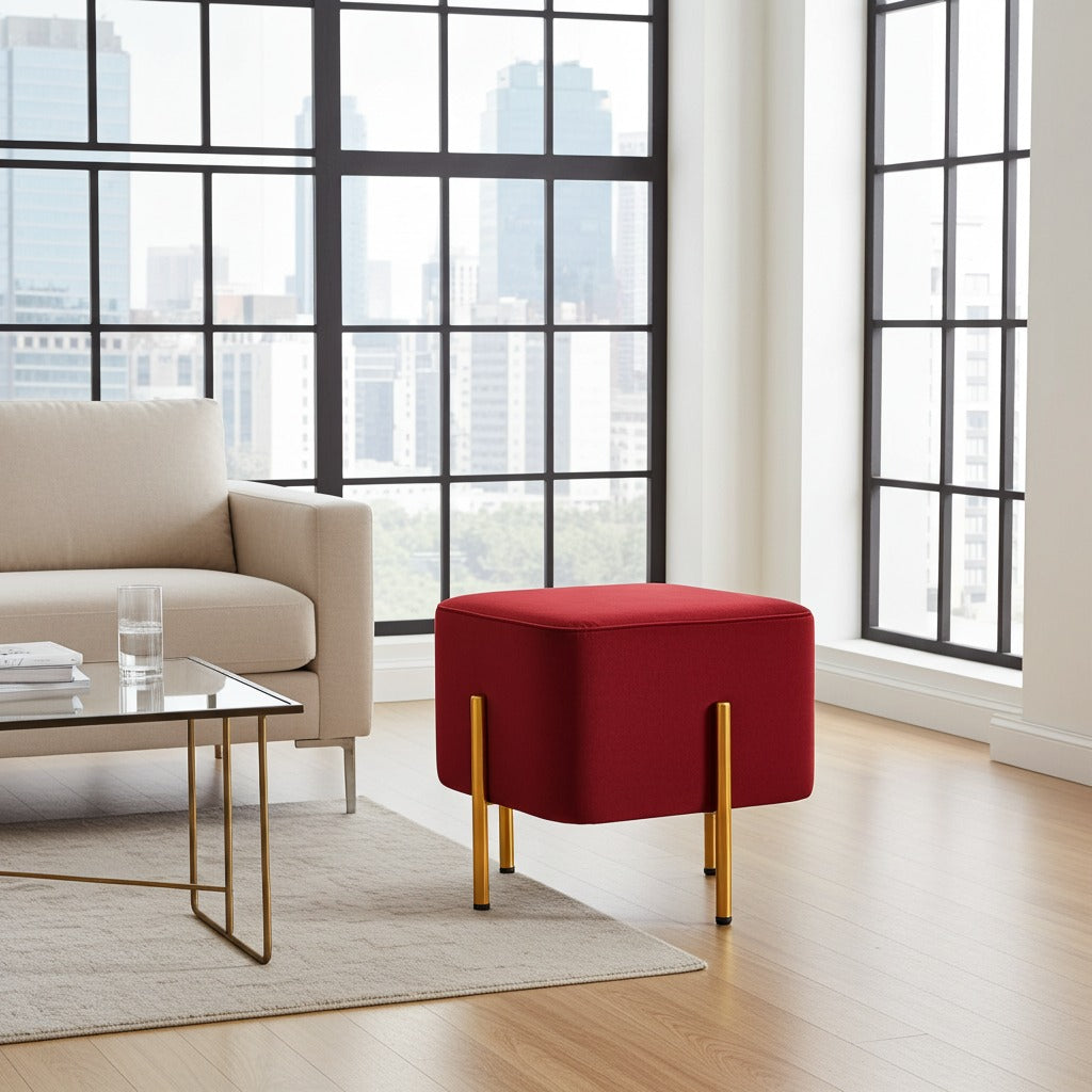 A square ottoman in deep red velvet with slim gold metal legs, styled in a modern living room next to a beige sofa and a glass coffee table.