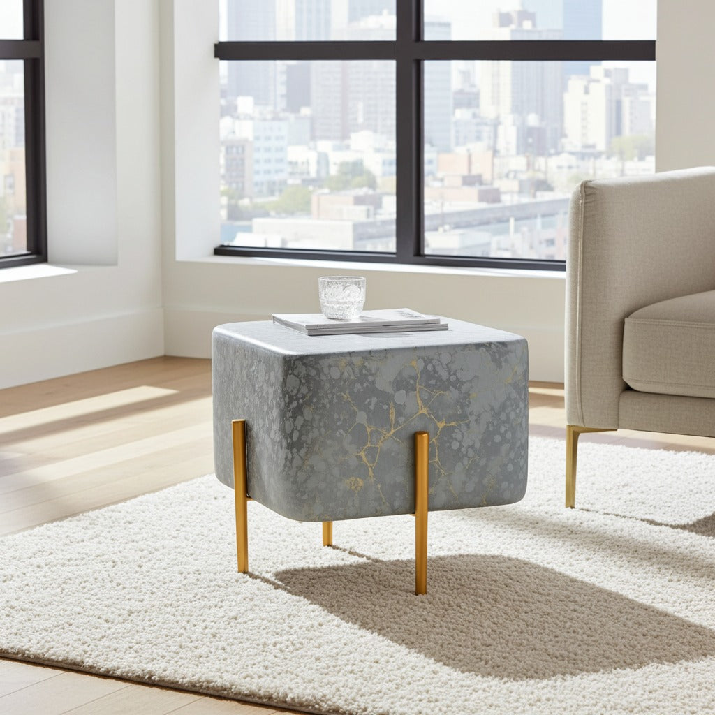 A modern square ottoman with a gray and white marbled patterned fabric top, supported by four gold-toned legs, styled in a bright living room next to a beige armchair.